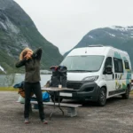 Person som tøyer nakke og rygg ved en bobil på rasteplass, illustrasjon til vondt i nakke bobil
