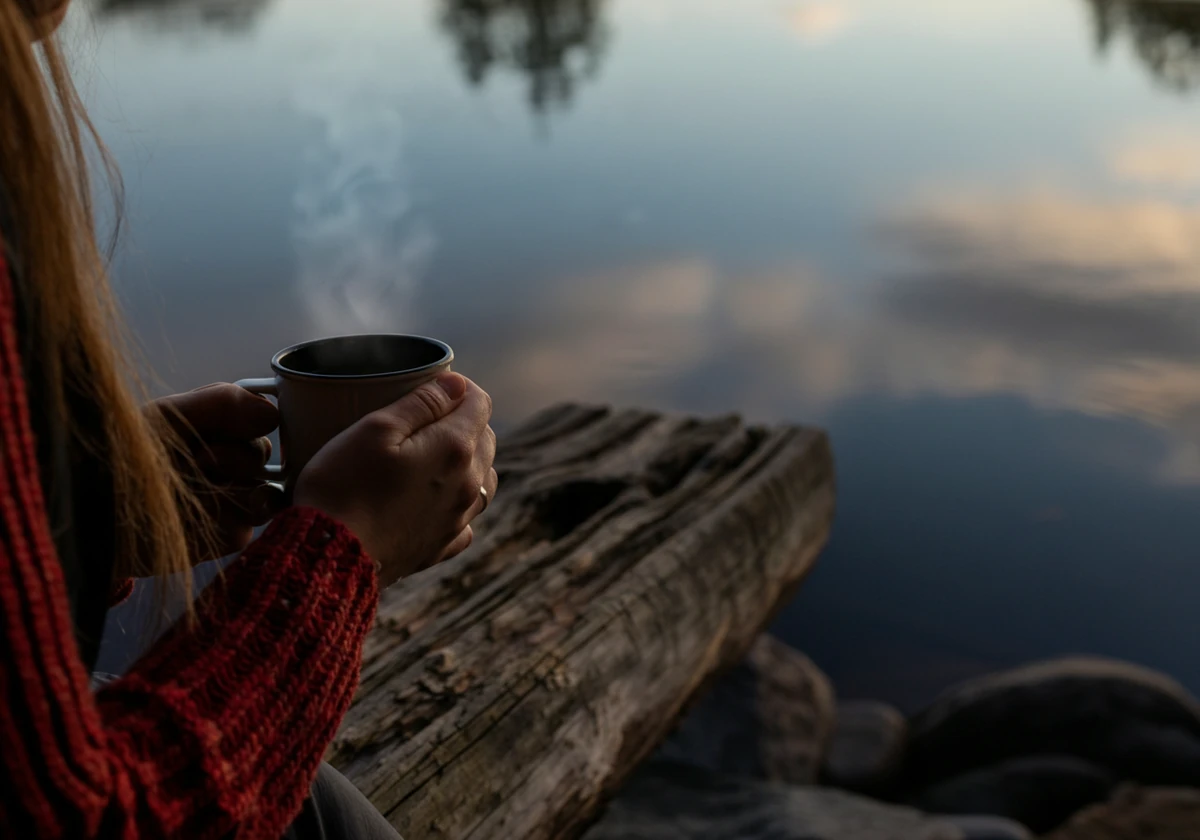 Person sitter med kaffekopp ved vannet på en teltplass tidlig om morgenen