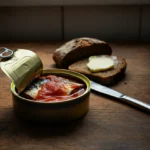 Åpnet boks med makrell i tomat på et trebord med skiver av grovt brød
