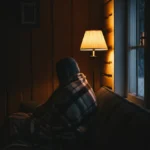 Person pakket inn i pledd i sofaen kjenner vinterdvale følelsen ved et mørkt vindu