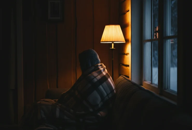 Person pakket inn i pledd i sofaen kjenner vinterdvale følelsen ved et mørkt vindu